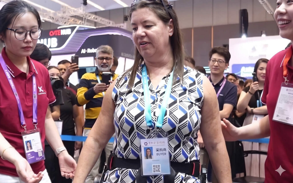 An Argentine woman with a muscle-wasting disease, long reliant on a wheelchair, stands and takes supported steps using a powered waist-assist exoskeleton developed by Hangzhou Taixi Intelligent Technology. The moment unfolds at the 139th Canton Fair in Guangzhou, China, as family members cry and the surrounding crowd applauds, showcasing how affordable Chinese assistive robotics is restoring mobility, dignity, and hope.